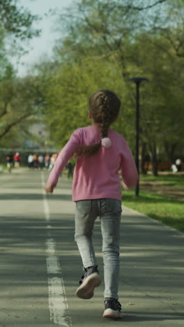playful little girl with braided fair hair runs along empty asphalt pedestrian road in spring park backside view. active sportive child has fun slow motion