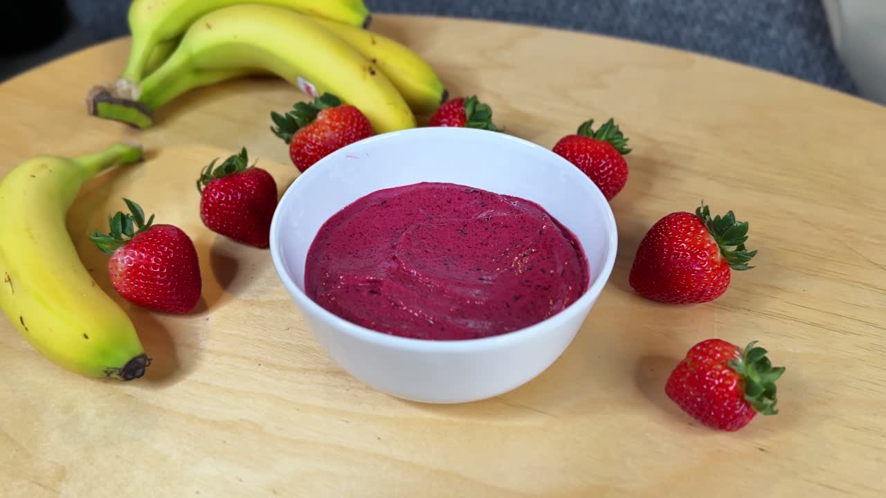 Una mano liscia un denso smoothie bowl all'açai con un cucchiaio, circondato da fragole fresche e banane: una composizione pulita e invitante, ravvicinata, perfetta per ricette, benessere e contenuti da bar