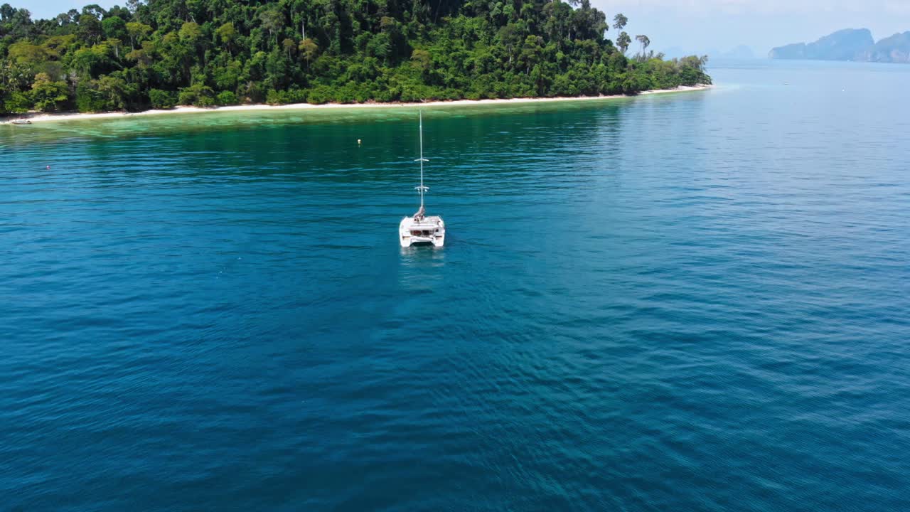 toma aérea de un catamarán nadando en el motor en el agua azul cristalina del mar de andaman con una isla tropical en el fondo