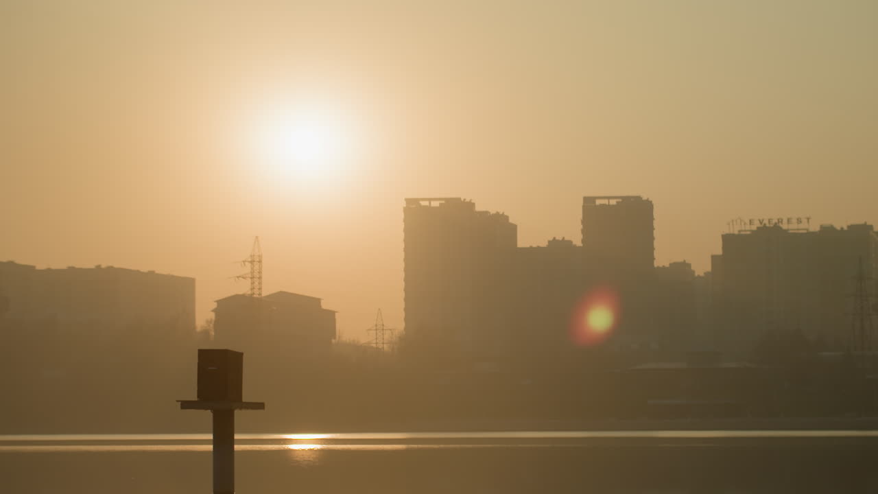Hazy sunrise city skyline reflected water, solitary post in foreground, amber glow over industrial silhouettes, calm waterfront atmosphere, cinematic early morning scene before commute