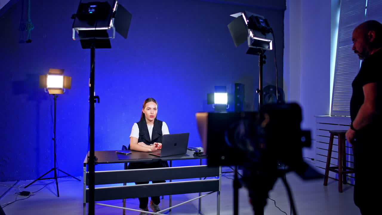 Caucasian girl sits talking in studio at the desk with laptop and phones on. Photographer comes up to the desk taking things from it.