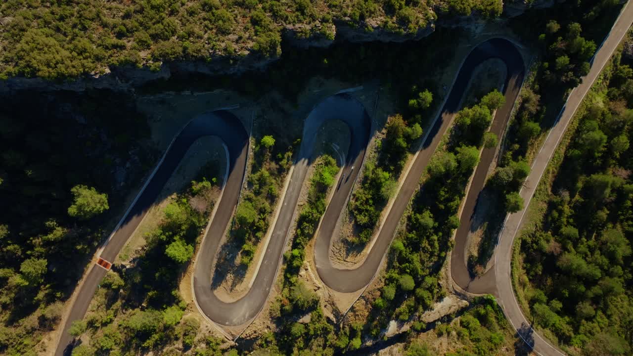 Uma foto estática e ampla, tirada por um drone, revela ciclistas percorrendo curvas fechadas e íngremes em ziguezague por uma montanha arborizada com vista para o desfiladeiro do rio Cinca, em Sobrarbe, com várias curvas visíveis.