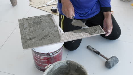 position a piece of ceramic tile on a bucket, a construction worker is plastering the mortar to the tile as he prepares to put it in place on the floor