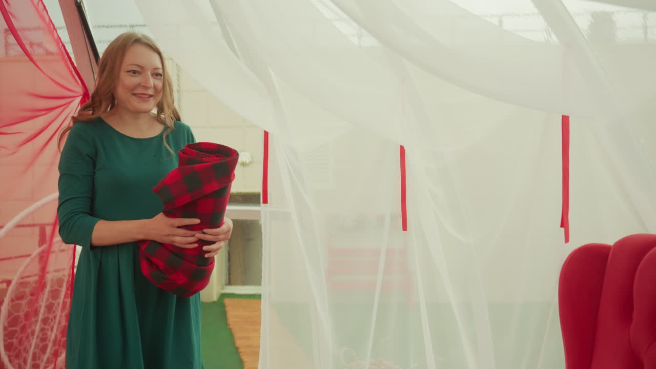 Una coppia condivide un plaid e sorrisi, una donna in abito verde offre una coperta a quadri a un uomo seduto in abito accanto a una poltrona rossa, tende leggere svolazzano, la calda luce del sole riempie l'interno, spontaneo