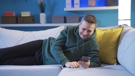 hombre feliz relajándose en el sofá en casa y navegando por las redes sociales en su teléfono inteligente, mirando las publicaciones.