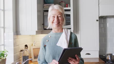 Portrait-of-happy-senior-caucasian-woman-standing-in-kitchen-and-using-tablet