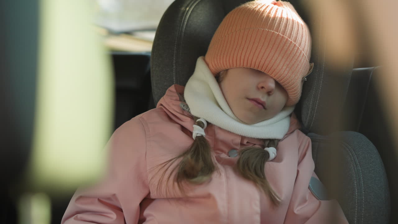 A young girl wearing a pink hat and scarf is sleeping in a car seat with her hat pulled down over her eyes
