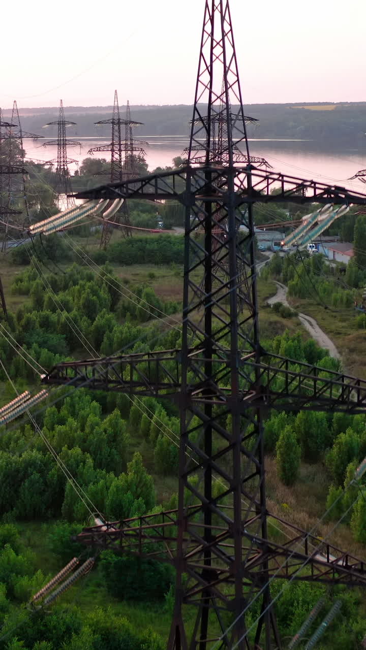 Structure of high-voltage lines outside the city at sunset. Trees and river. Camera motion back. Aerial view. Vertical video