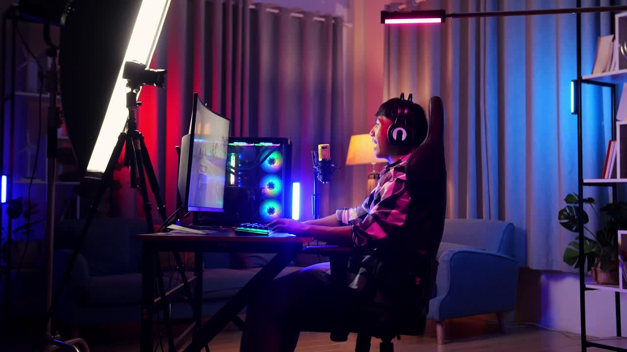 vista lateral de un niño asiático jugando a un juego en una computadora personal, agitando la mano hablando con los participantes en el micrófono. videojuego de transmisión en vivo, escritorio iluminado por luz de tira led rgb