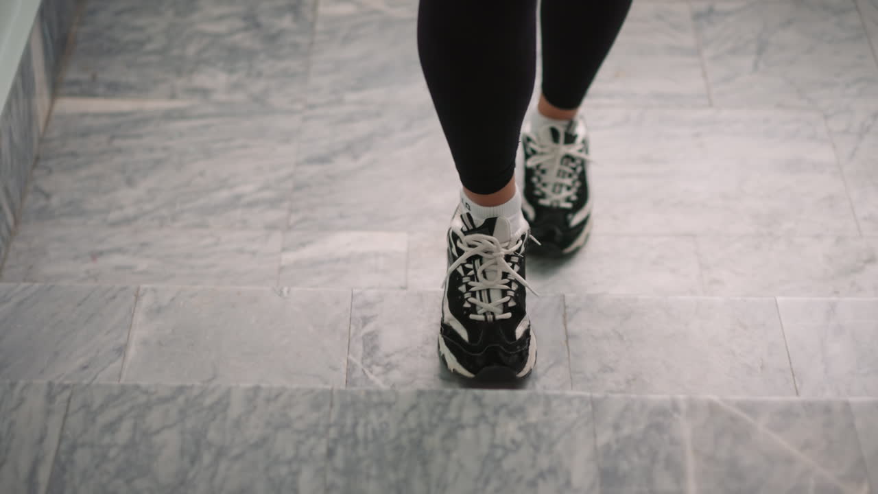 Close view of person legs in black white sneakers walking up marble staircase, steady upward motion with focus on steps, laces and socks, indoor scene suggesting fitness, routine commute