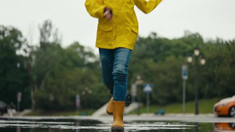 Ein-Glückliches-Blondes-Mädchen-In-Einer-Gelben-Jacke-In-Gummistiefeln-Läuft-Durch-Eine-Pfütze-Und-Spritzt-Nach-Dem-Regen-Wasser-An-Die-Seiten-Im-Park