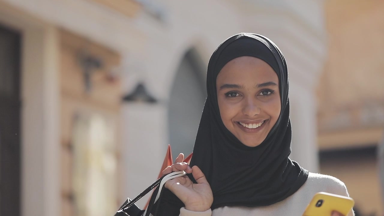 Giovane donna con hijab che usa lo smartphone e porta borse della spesa