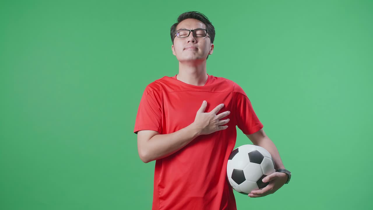 Asian Man With A Ball Putting Hand On His Heart To Respect The National Flag And Anthem While Cheering Soccer On Green Screen Background