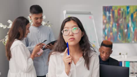 brunette with glasses is sitting on desk in company office wearing plaid skirt white shirt, she is putting pen to cheek in hand holding notebook, deep contemplation, reverie, looking for an idea