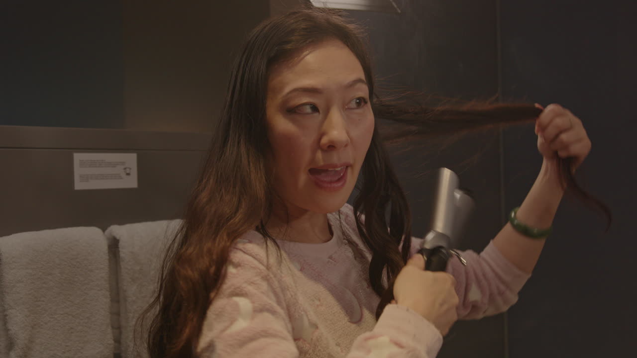 Close up of an attractive Asian woman curling her hair and doing a little dance in a hotel bathroom. Smoke rises from her hair when the girl releases the curling iron