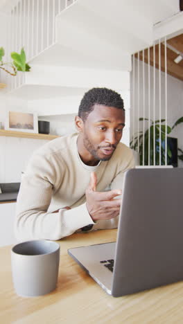 vertical video of african american man leaning on countertop in kitchen, using laptop
