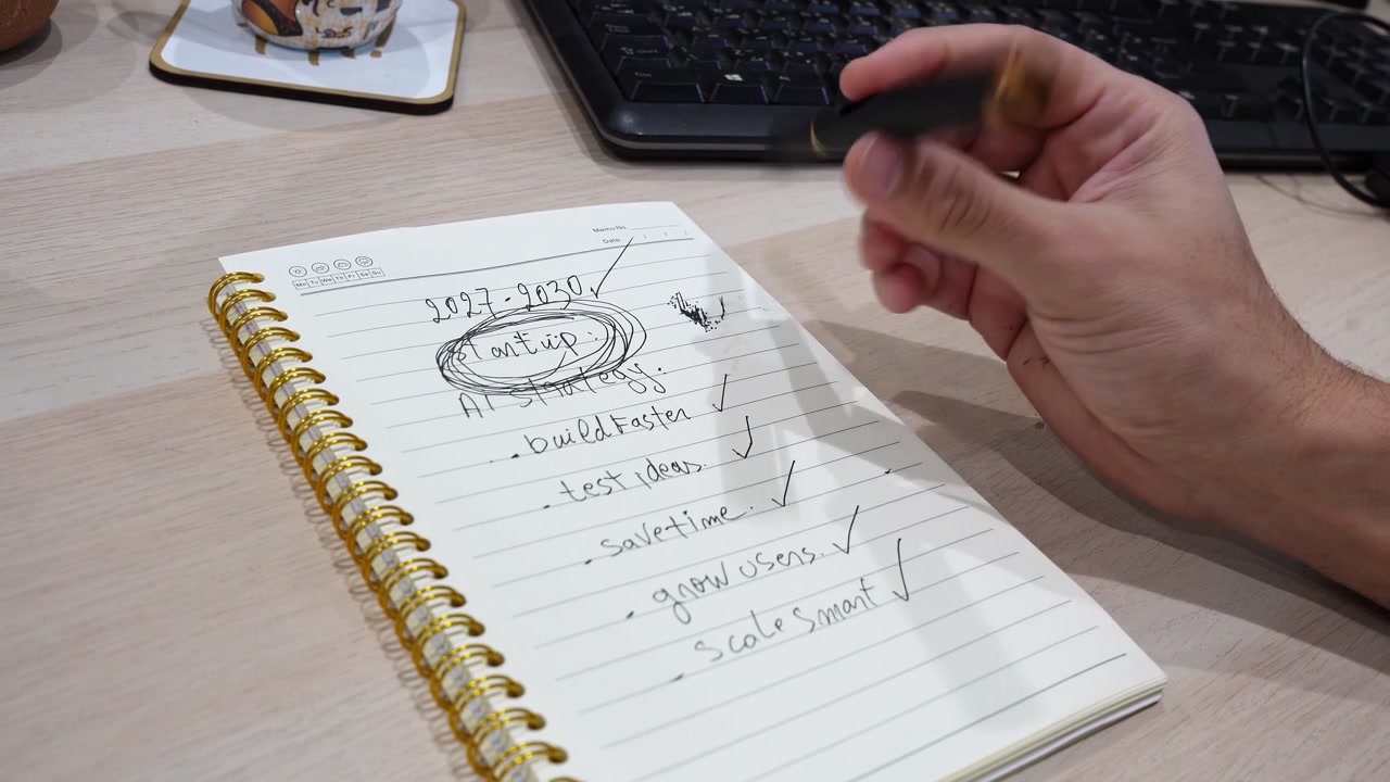 Tampilan dekat tangan yang memutar pena di atas meja kantor. Buku catatan menunjukkan tujuan strategi AI yang ditulis tangan untuk startup 2027-2030, termasuk membangun lebih cepat, menguji ide, dan meningkatkan skala dengan cerdas