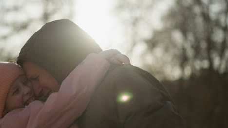 a little girl in a pink cap and jacket being lifted up playfully by a man wearing a brown jacket and blue jeans as they laugh joyfully. the warm sunlight highlights their happy expressions