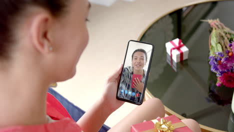 Biracial-woman-holding-smartphone-with-biracial-woman-with-gift-on-screen-with-gift-on-desk