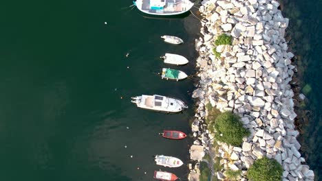 Vista-Aérea-De-Pájaro-De-Barcos-Pesqueros-Atracados-En-Un-Pequeño-Puerto-De-Piedra,-Kavala-Perigiali,-Grecia