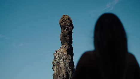 silhouetted woman gazes at ilhéus da ribeira da janela’s vertical sea stack, its rugged peak framed against a wide blue sky in peaceful stillness.