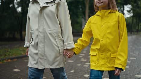 Close-up-a-blonde-teenage-girl-in-a-yellow-jacket-walks-hand-in-hand-with-her-mother-along-an-alley-in-the-park-after-the-rain