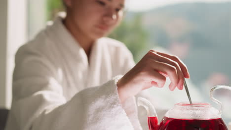 lady stirs tea in teapot sitting at table closeup. relaxed woman in bathrobe brews hot hibiscus tea sitting by window. concept of weekend morning