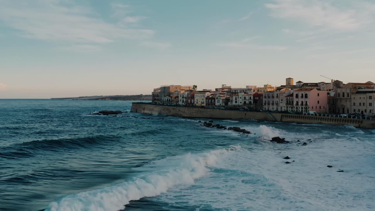 Flying low over the waves in the sea near historic medieval town in Ortigia island at sunset. Waves crashing on the shore.