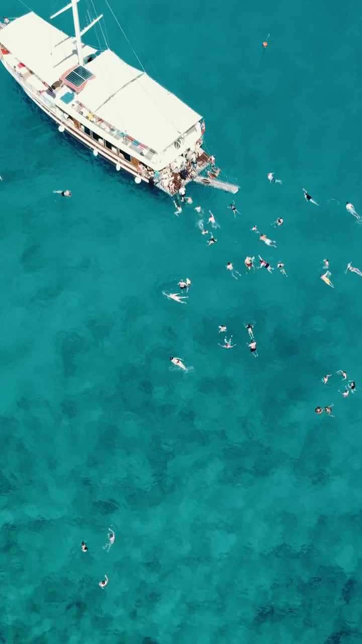Aerial drone view showing leisure boat surrounded by swimmers and snorkelers in clear blue sea near Kekova coast, perfect for Social Media travel posts and ocean lifestyle scenes