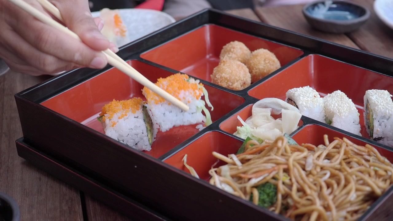 Person, die mit Essstäbchen eine Sushi-Rolle aus einer Bento-Box nimmt