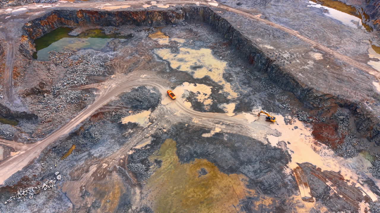 Aerial shot of excavator and truck working in stone quarry. High angle view of yellow excavator loading material into a dump truck near water puddles in a granite pit