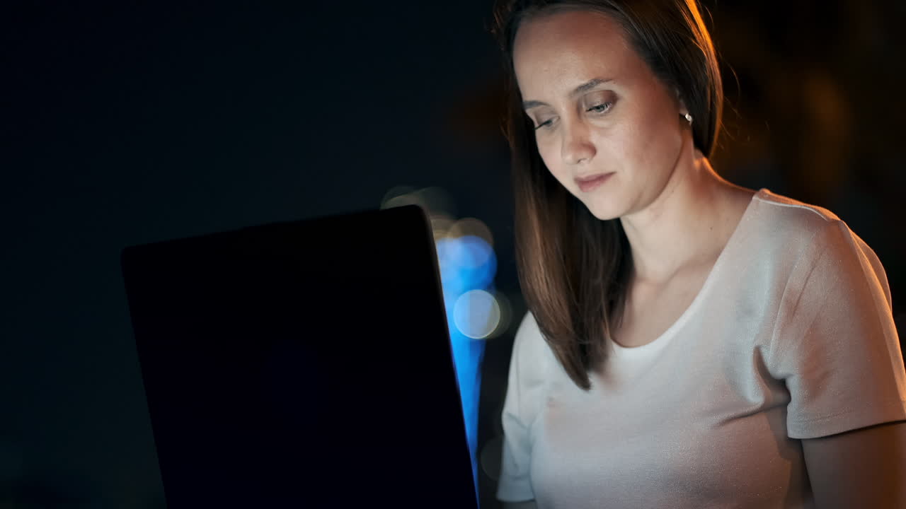 A woman software developer sits in a Park on a summer night in the city and writes code looking at a laptop screen. Print the article on a laptop. Blogger replies to posts
