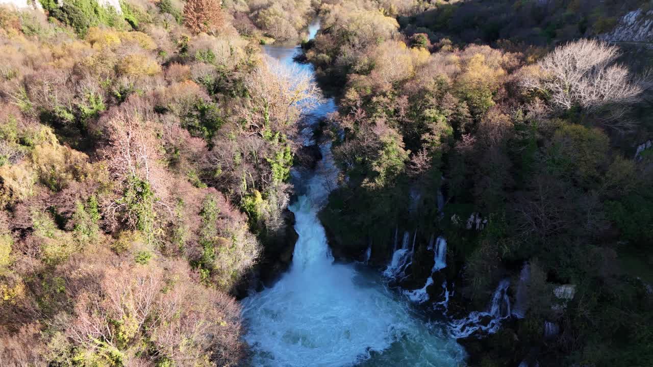 A drone aerial captures Bilusića Buk waterfall along the Krka River in Croatia. The surrounding forest displays vibrant autumn colors, with sunlit and shadowed areas creating depth and texture