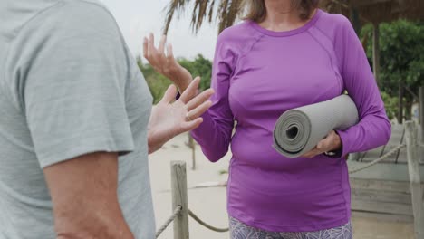 Happy-senior-caucasian-couple-holding-yoga-mats-and-talking-on-beach,-in-slow-motion