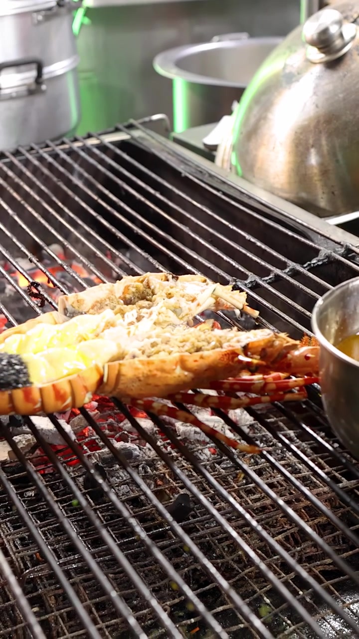 Chef con guanti spennella la salsa su un'aragosta divisa a metà su una griglia calda, illuminazione vivace di un mercato di street food