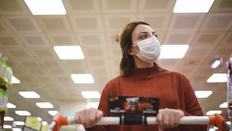 beautiful woman wearing protective disposable medical mask and fashionable clothes uses shopping cart while shopping in supermarket