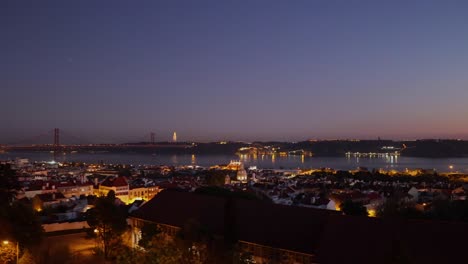 Aerial-view-of-Alfama-in-Lisbon-with-some-cars-going-home-on-the-bridge-25-de-Abril-and-Cristo-Rei-in-the-brightly-lit-background,-Lisbon,-Portugal-at-night-from-restelo