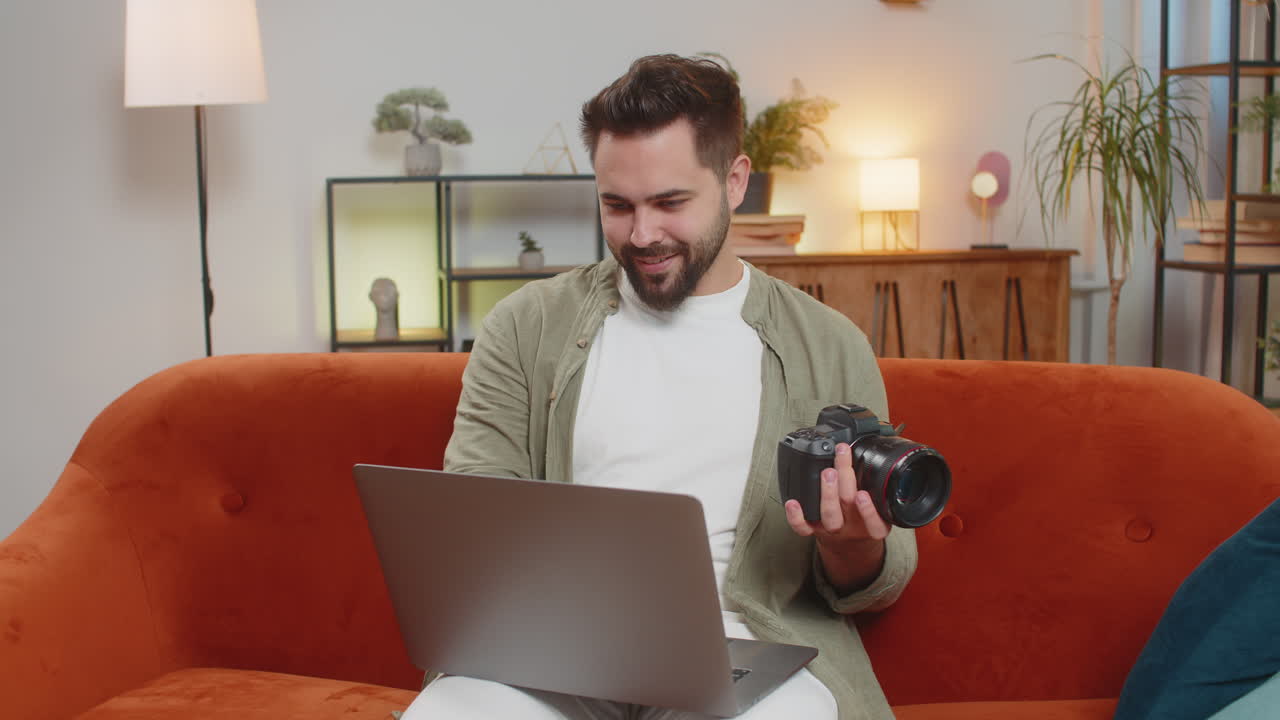 Happy man photographer with laptop holding digital camera looking at screen choosing photos at home
