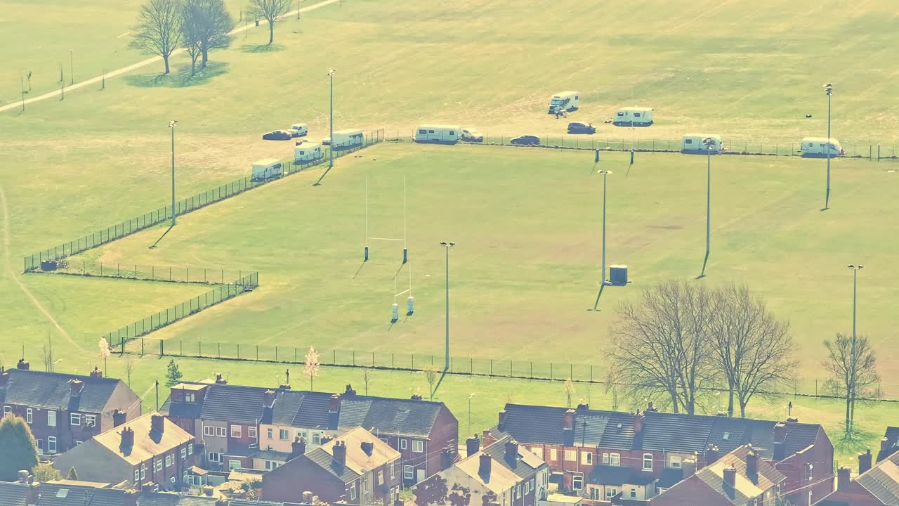 Aerial view of Rotherham suburb featuring residential houses, a playing field with rugby posts, caravans parked on the field, and trees, all bathed in natural daylight