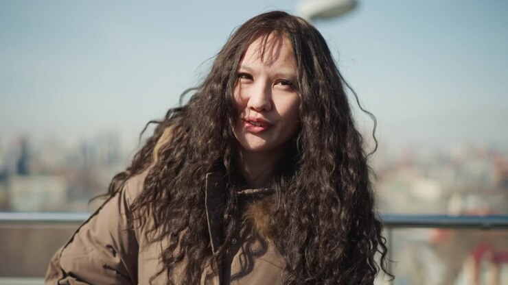 Donna con lunghi capelli ricci indossa una giacca marrone e si trova davanti a uno sfondo urbano sfocato. La consistenza dei suoi capelli scuri ondulati è visibile insieme ai dettagli del suo abbigliamento casual