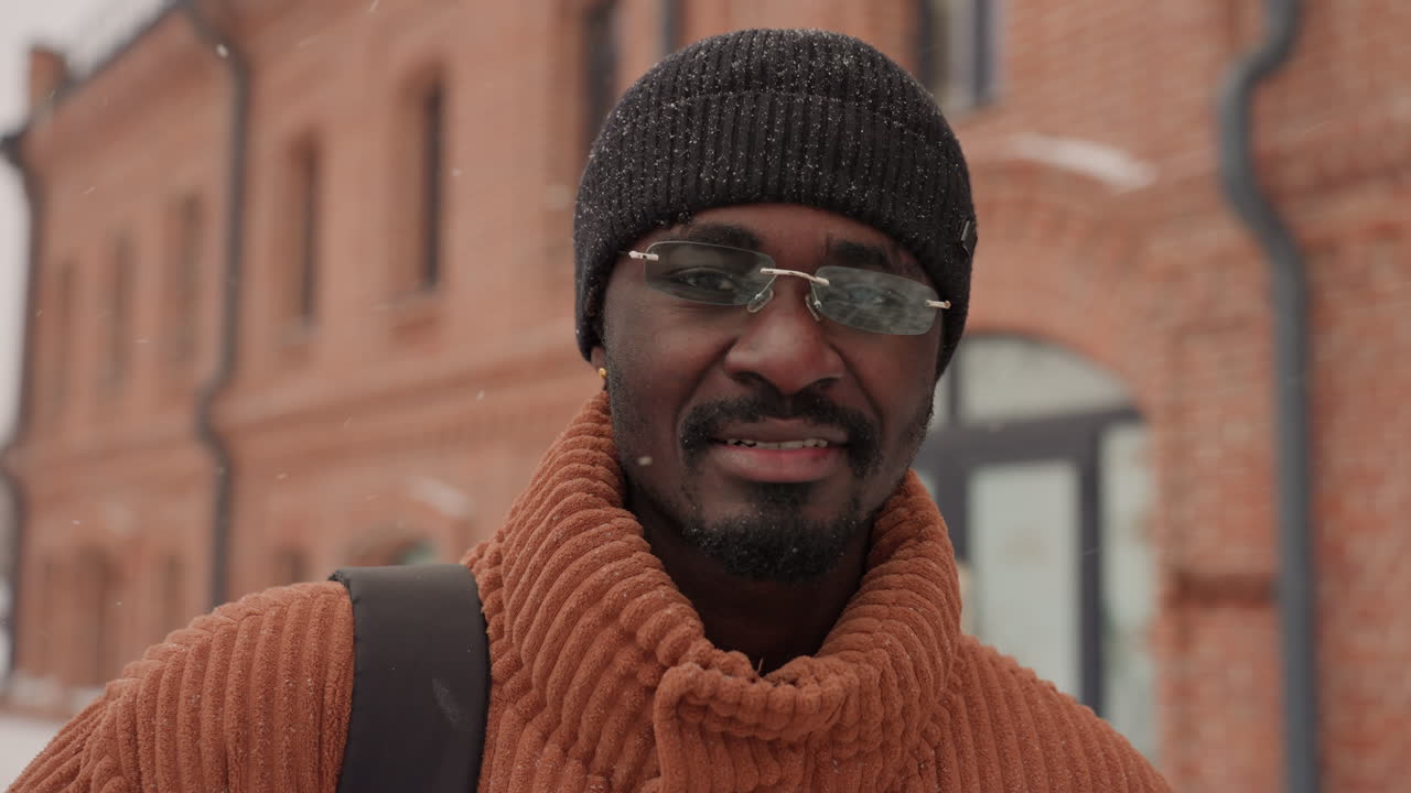 Uomo sorridente vicino a un edificio in mattoni in una città invernale, maglione caldo color ruggine e cappello di lana con fiocchi di neve, tracolla dello zaino visibile, sguardo spontaneo verso la fotocamera vicino a un ingresso e a pedoni, ruoli includono turista, locale.