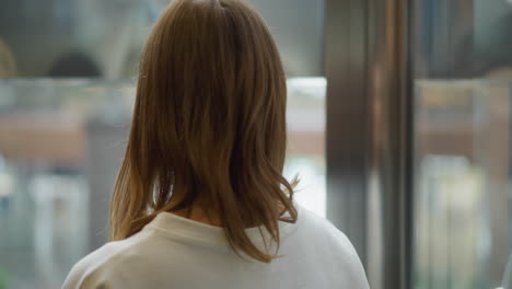 back view of a young woman inside a moving elevator with soft natural lighting, offering a blurred glimpse of the urban environment through glass panels