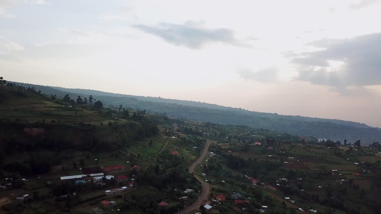 Drone shot over Kapchorwa, Eastern Uganda, showing rolling green hills, a winding road, scattered homes, and distant sunset clouds over the Elgon slopes during early evening light