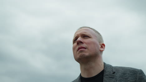 a close-up view of an aged white man in a grey suit and black polo shirt, looking intently into the distance with a determined expression, with an overcast sky