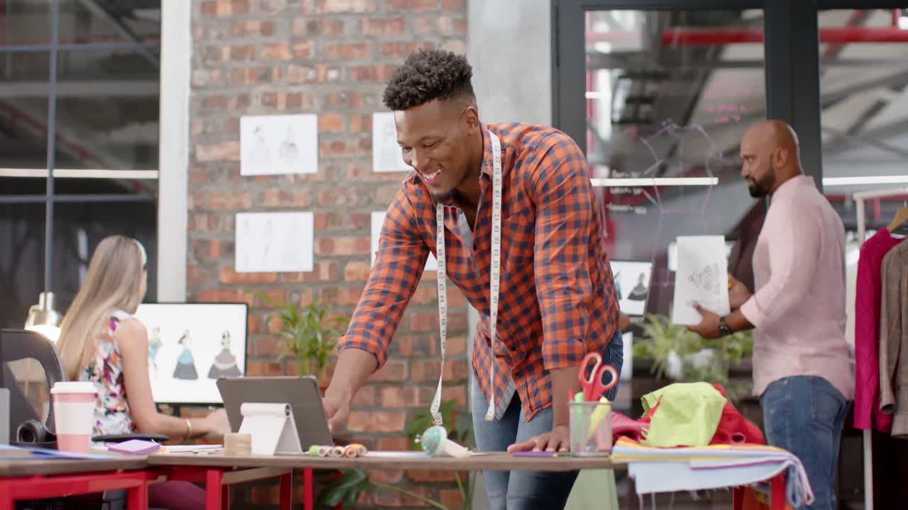 retrato de un feliz diseñador masculino biracial usando una tableta y riendo en un estudio de moda, en cámara lenta