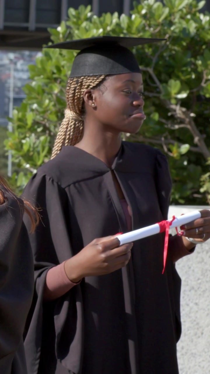 Vertical video: Turning graduate in gown adjusting, checking diploma with ribbon at campus ceremony