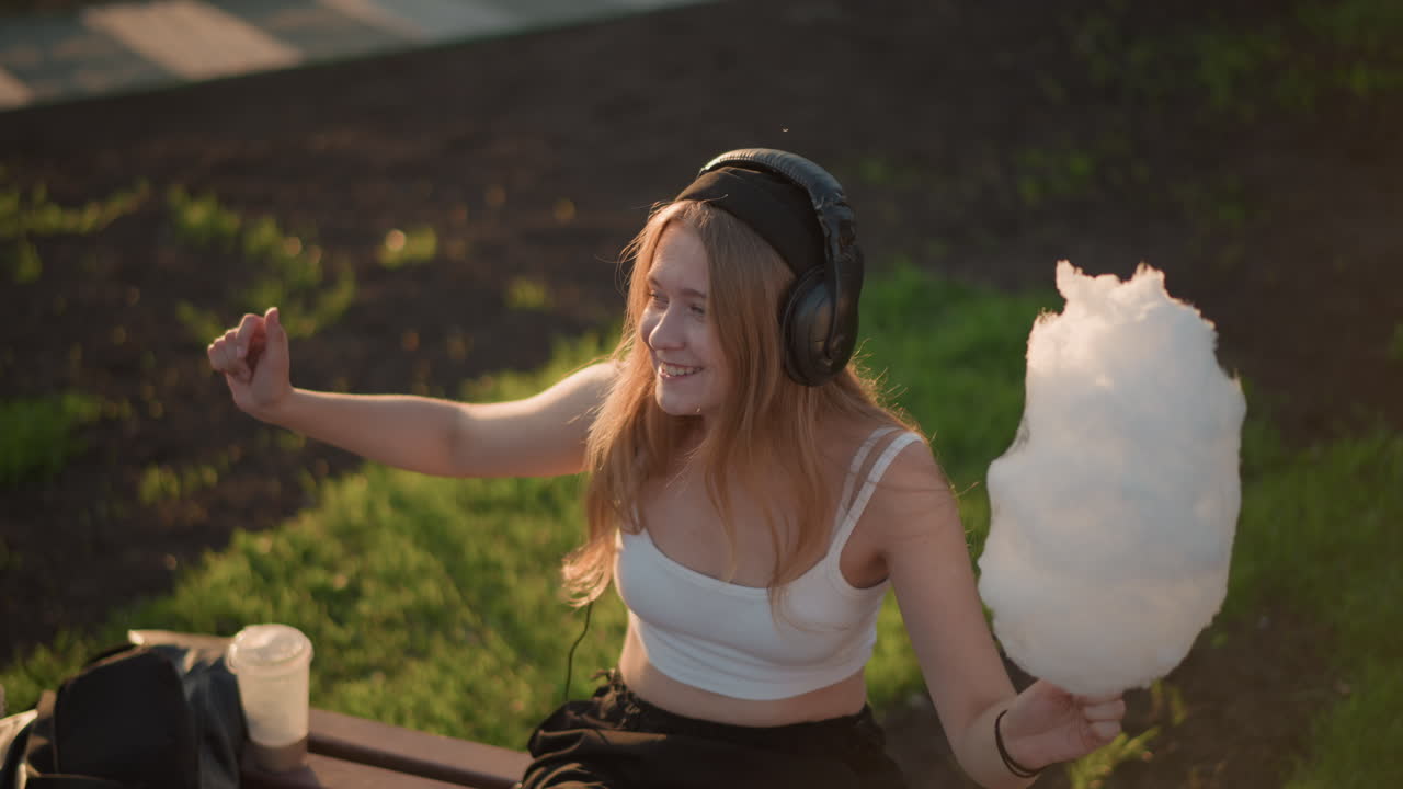 young woman dancing on bench with cotton candy and headphones, sunset sunlight casting warm glow rhythmic arm movements and laughter, upbeat energy and urban park atmosphere, playful munching
