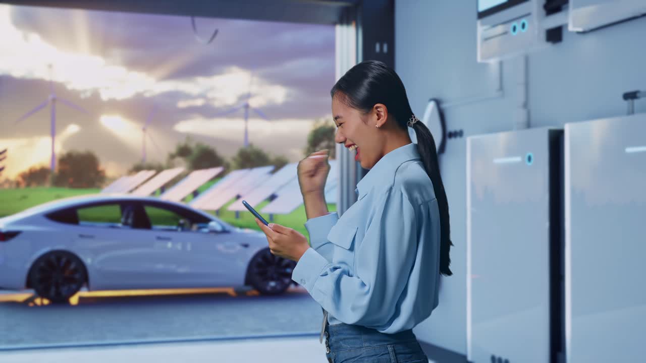 Side View Of Asian Female Professional Worker With Her Smartphone With Home Energy Storage System In a Modern Garage, She Raises Her Fist Up With Screaming Goal After Check On The Smartphone