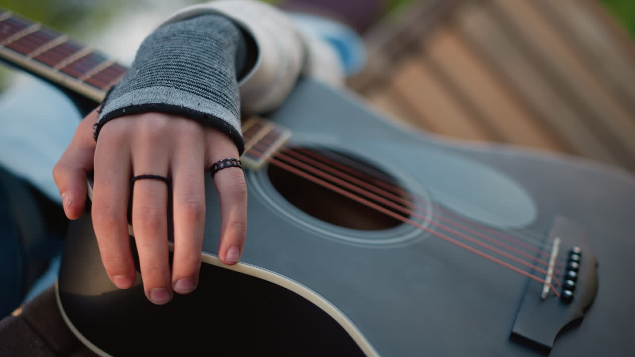 Primo piano di una mano appoggiata su una chitarra acustica, dita adornate con anelli e braccialetto, polsino di un maglione testurizzato, foro e ponte dettagliati, pratica intima di fingerstyle, atmosfera da esibizione in un parco urbano