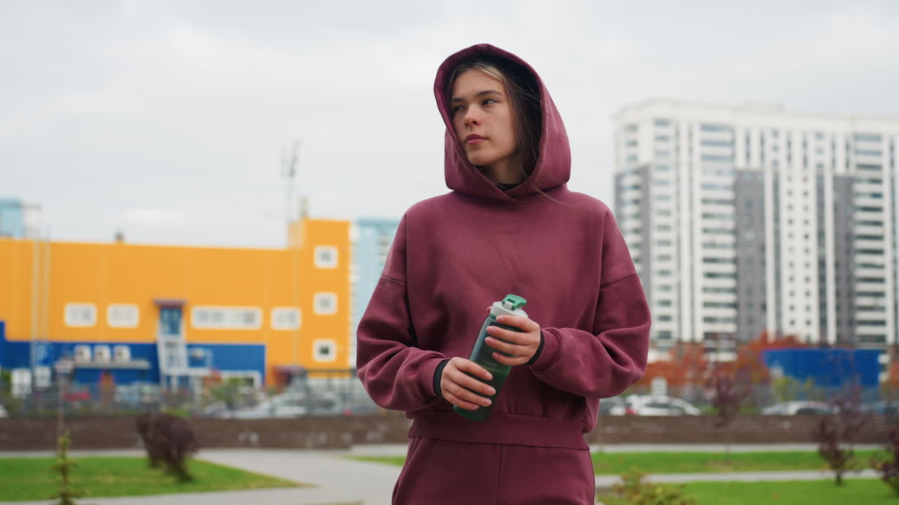 Fit woman walking on urban pavement clutching green water bottle while passing red spring flowers and tall modern apartment buildings under cloudy sky serene fitness lifestyle portrait